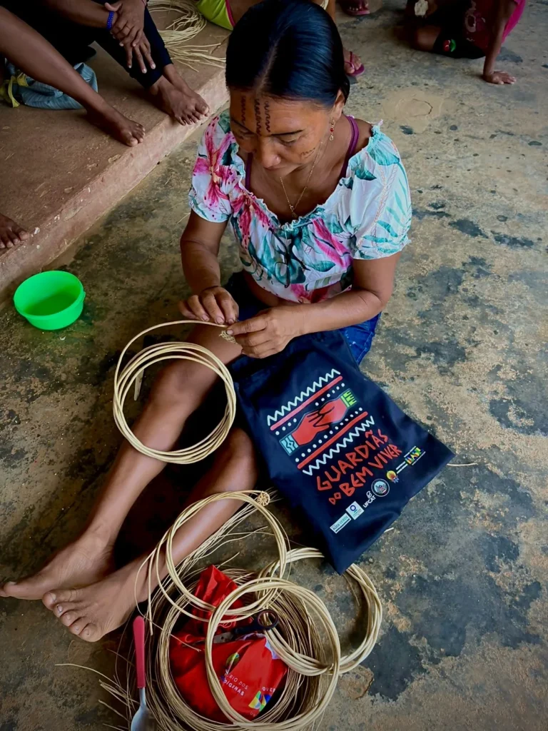 A Yanomami woman weaving fibers, seated next to a Guardiãs do Bem Viver bag