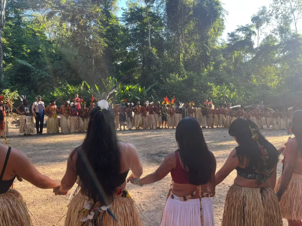 Indigenous women holding hands in a circle in a forest clearing, facing a line of participants